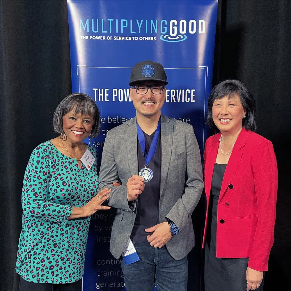 A man wearing a medal poses with two women in front of a blue Multiplying Good banner and black curtains.