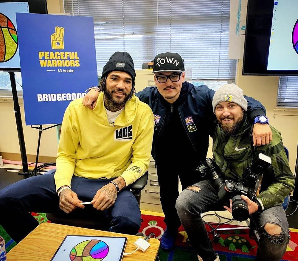 Three men pose together in an art-filled room, with the man on the right holding cameras.