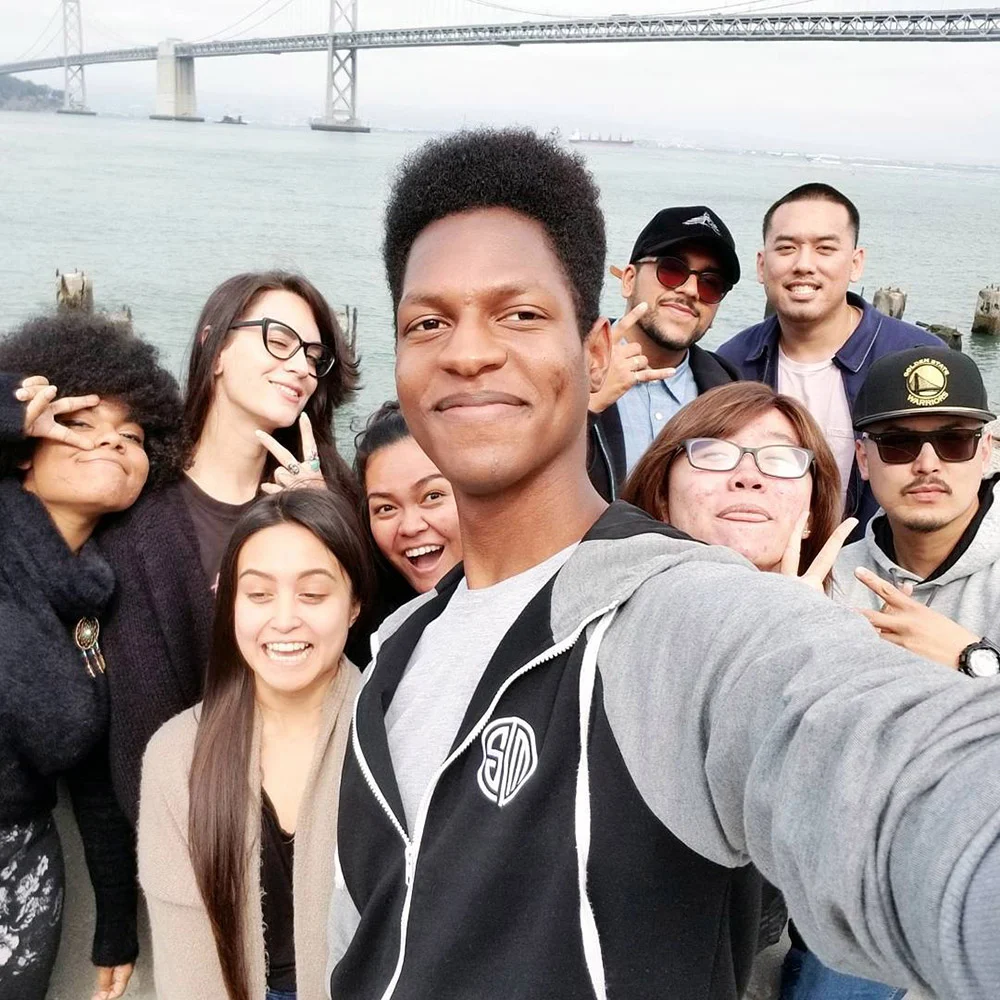 Eight friends crowd together for a waterfront selfie with the Bay Bridge behind them.
