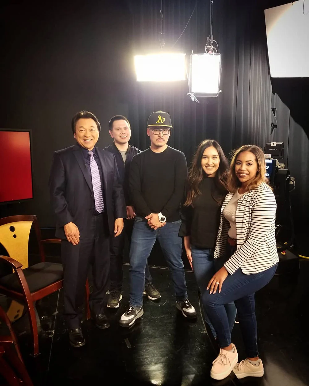 Five adults pose together in a studio with bright overhead lights, black curtains and wooden chairs.