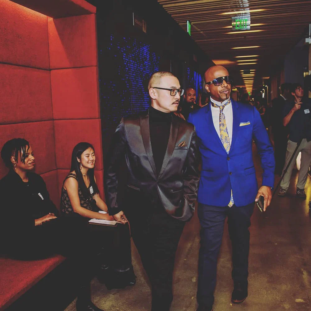 Two men in suits stand beside seated guests in a dim hallway with red padded walls.