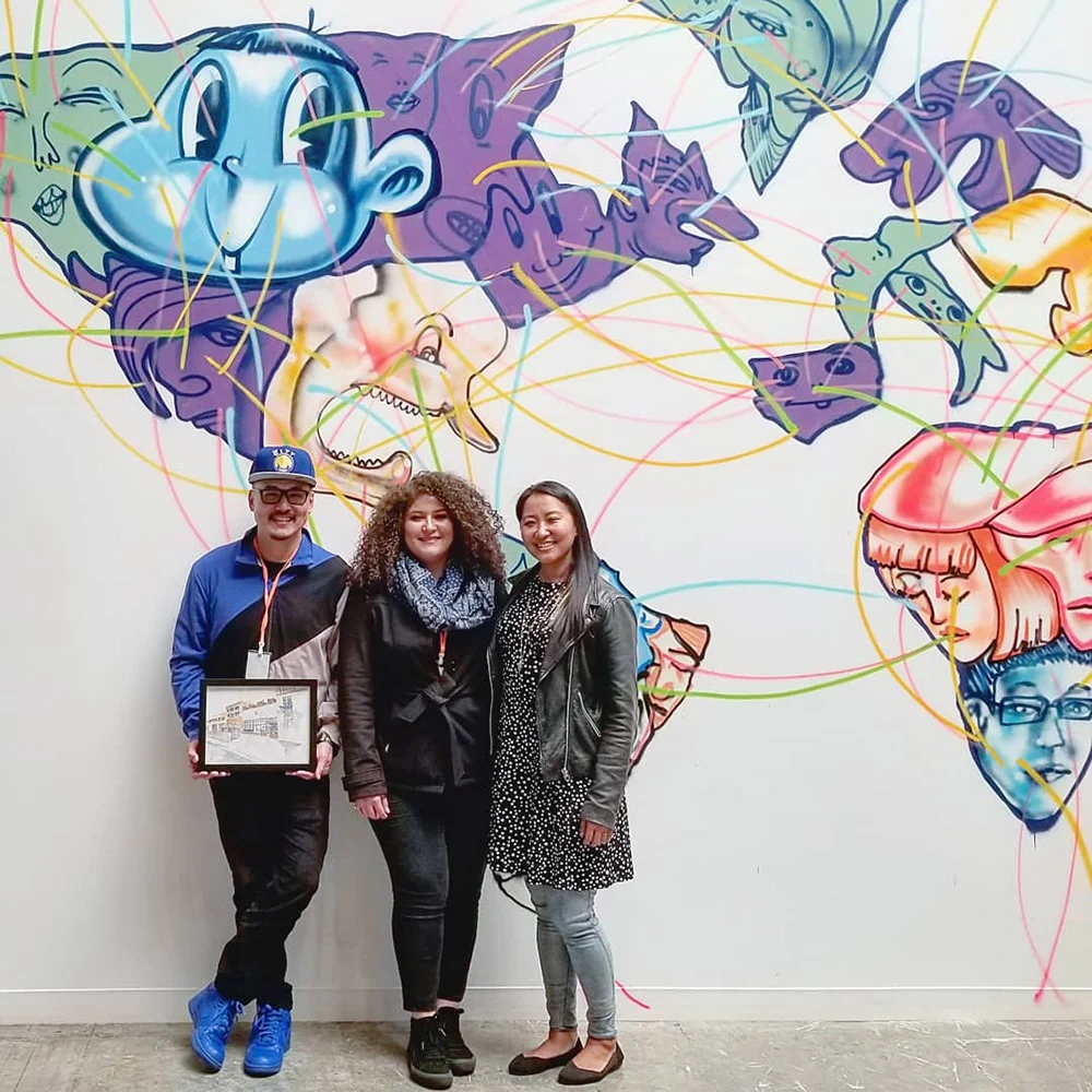 Three people pose in front of a colorful mural of cartoon faces, with one holding a framed piece.