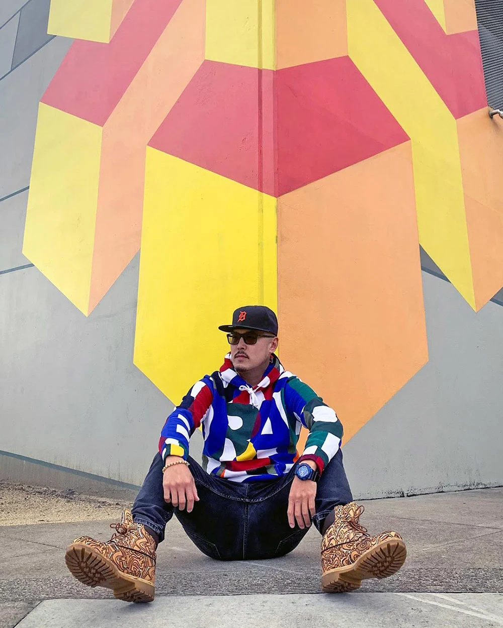 A man in a colorful hoodie, cap and boots sits on a sidewalk in front of a yellow, orange and red geometric mural.