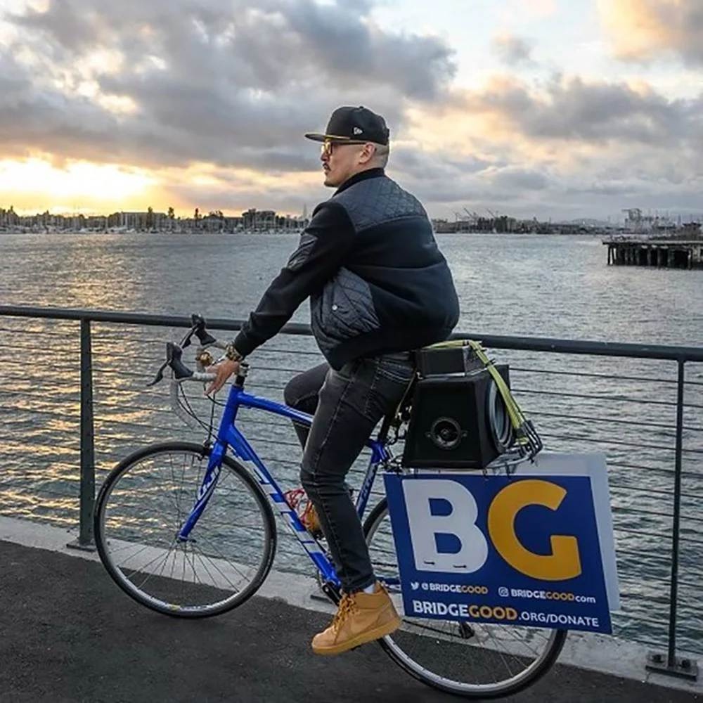 A person sits on a blue bicycle carrying a speaker and BRIDGEGOOD sign beside a waterfront railing at sunset.