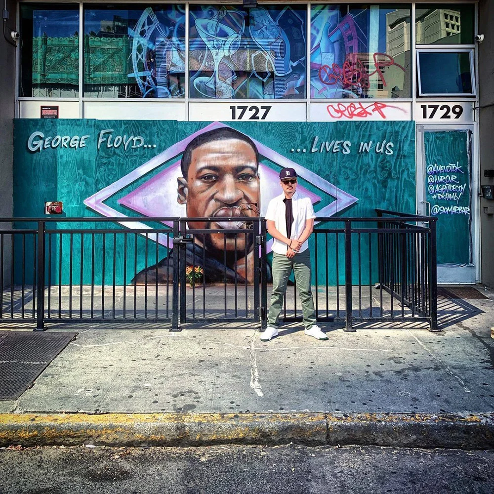 A person stands on the sidewalk in front of a George Floyd mural, behind a black metal railing.