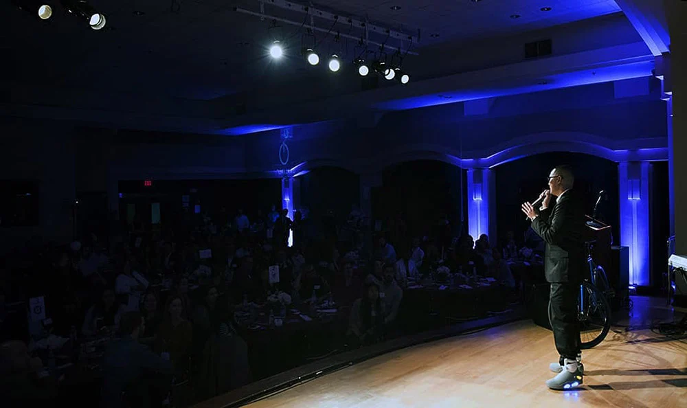A speaker holds a microphone on a wooden stage facing a seated audience in a blue-lit room.