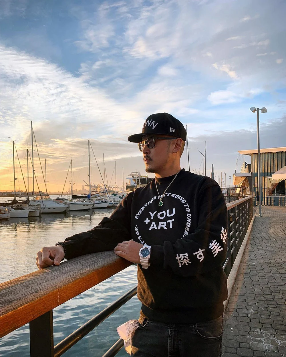 A man in a black cap, sunglasses and sweatshirt leans on a waterfront railing beside docked sailboats.