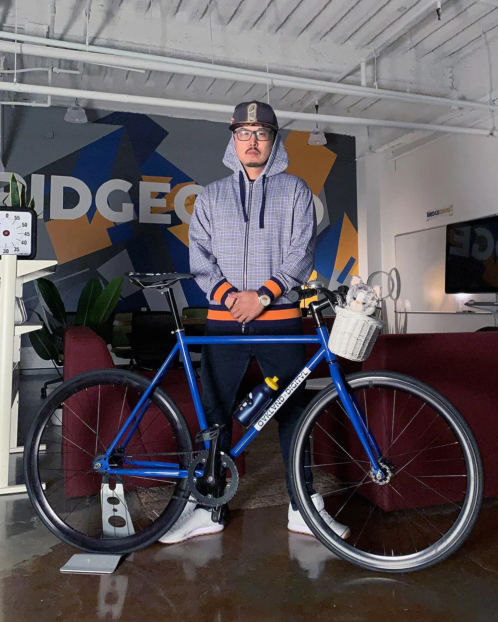 A person in a hoodie and cap stands behind a blue bicycle in a room with a colorful mural and red couch.