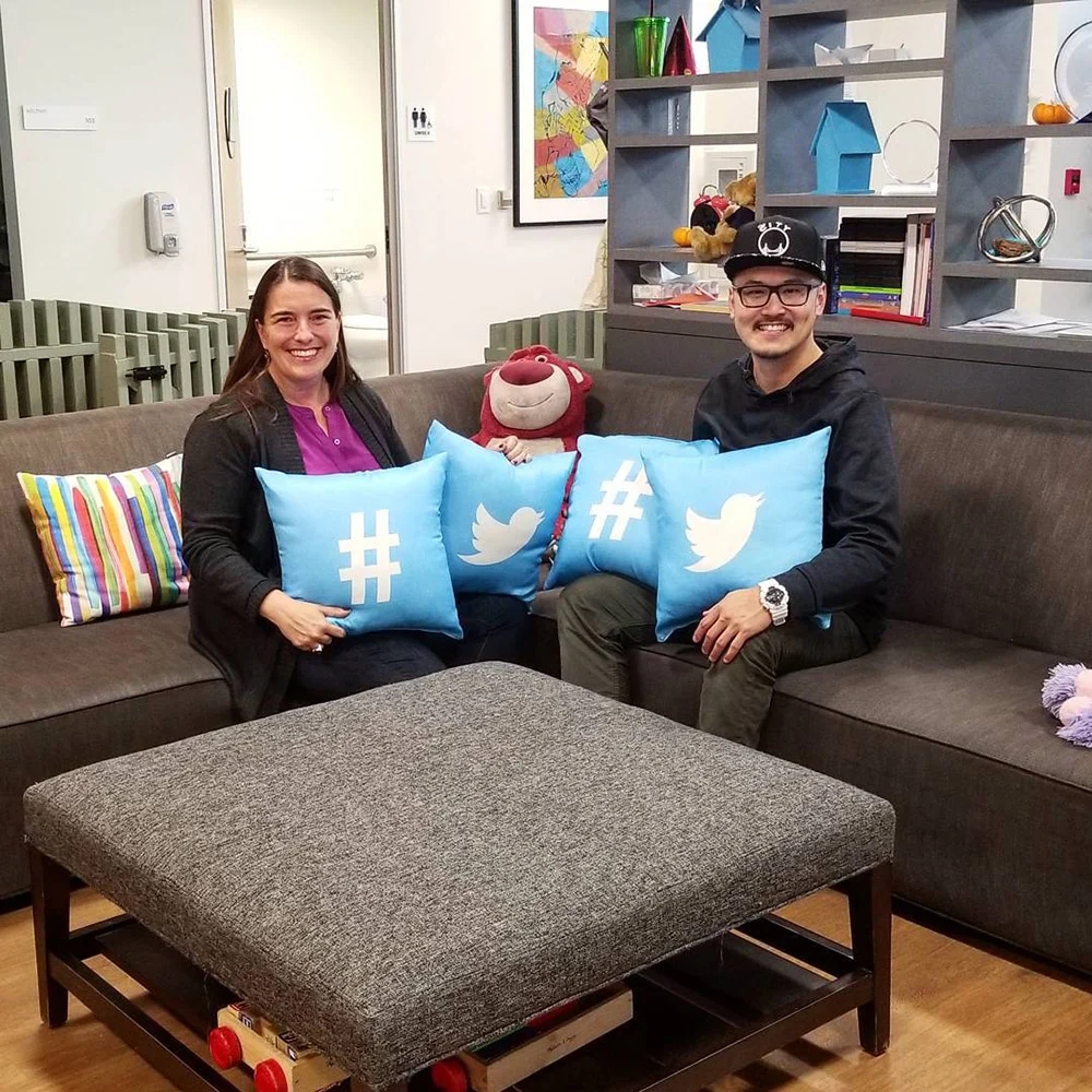 Two people sit on an office couch holding blue pillows with Twitter birds and hashtags, with a plush bear between them.