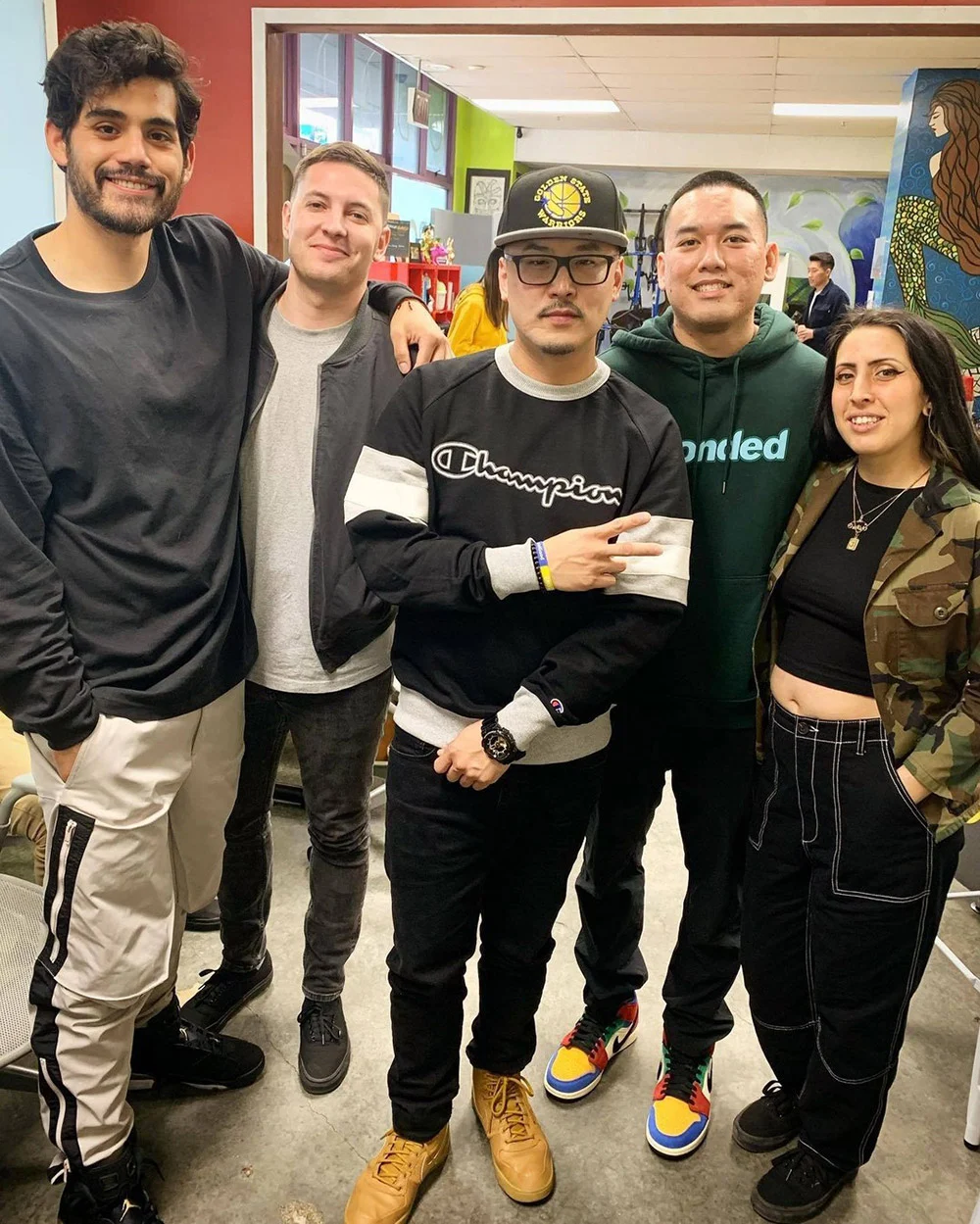Five people stand close together and pose in a colorful indoor space with murals and other people behind them.
