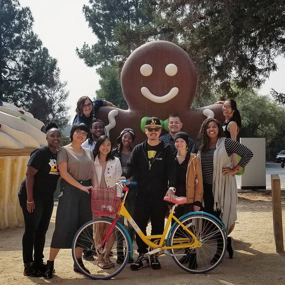 A group poses with a colorful bicycle in front of a giant gingerbread figure and trees.