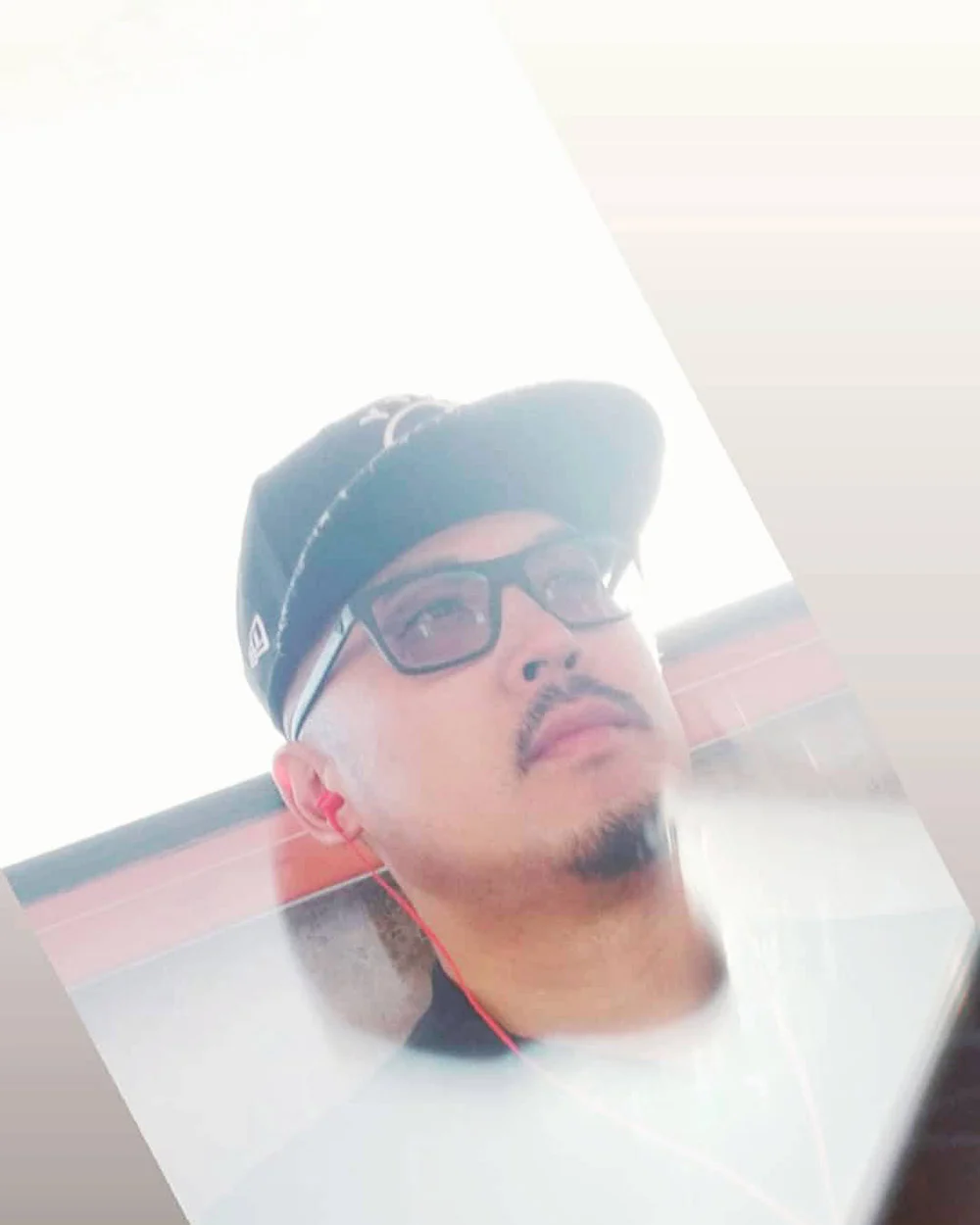 A man in a black cap, glasses and red wired earbuds looks upward beside a bright window.