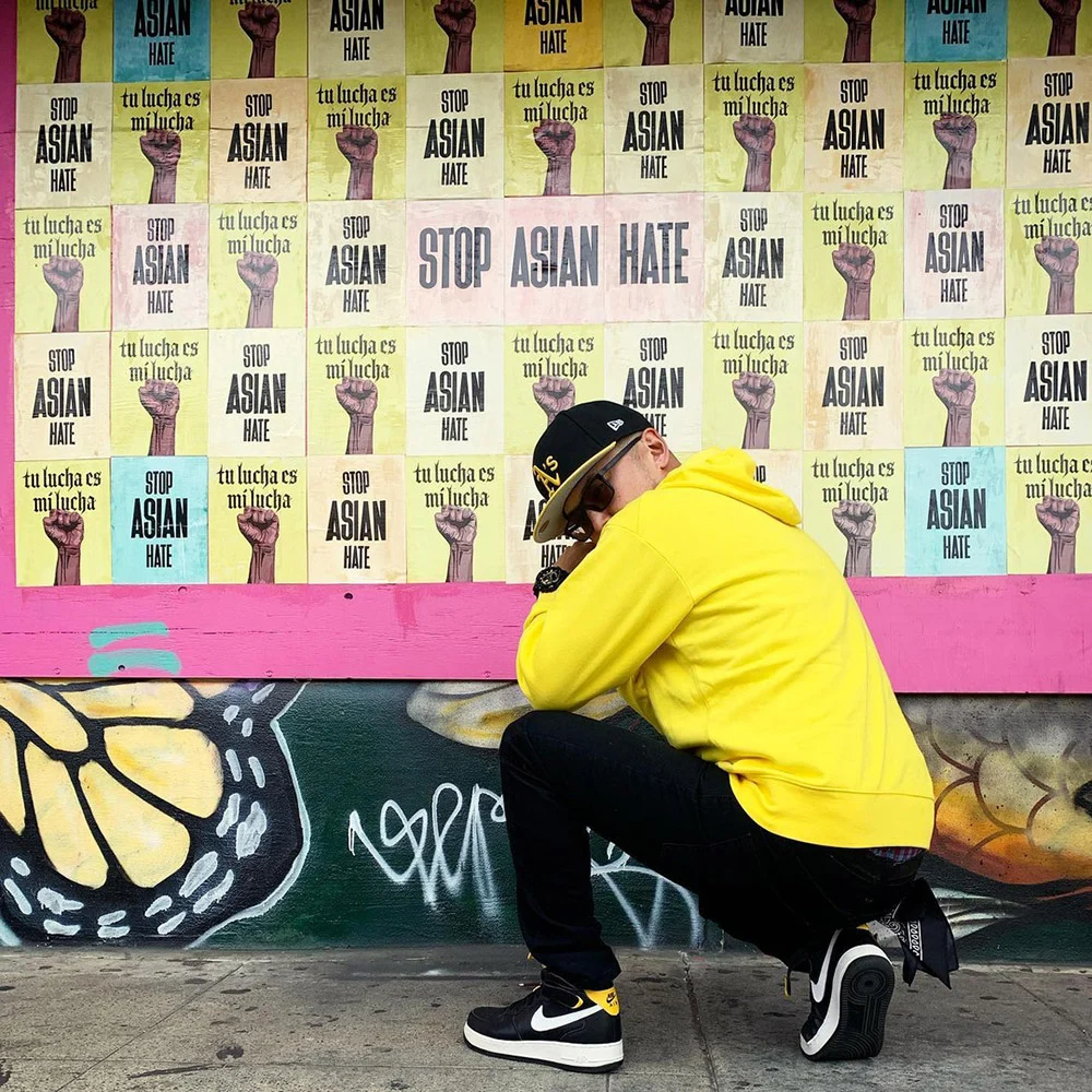 A person in a yellow hoodie crouches beside a mural beneath Stop Asian Hate posters with raised fists.