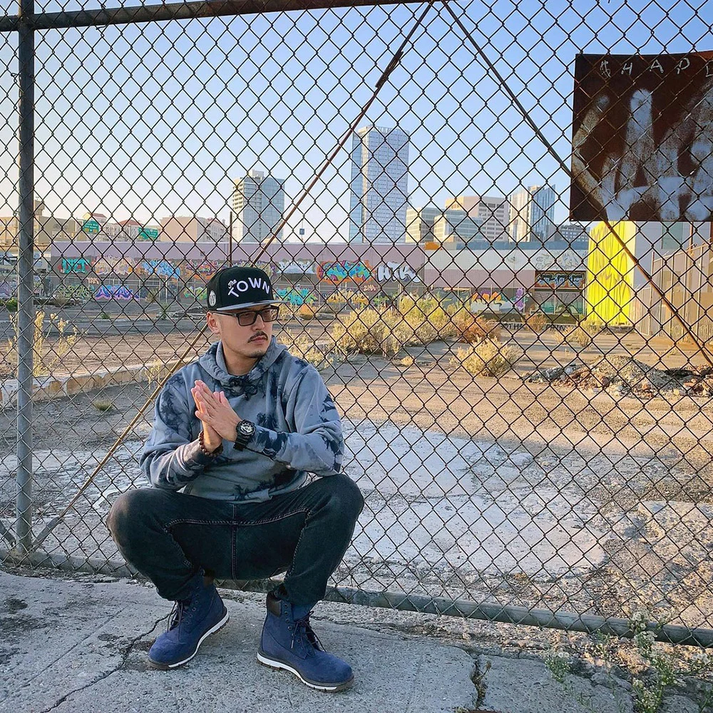 A man in a cap and sunglasses crouches with his palms together beside a chain-link fence overlooking a vacant lot.