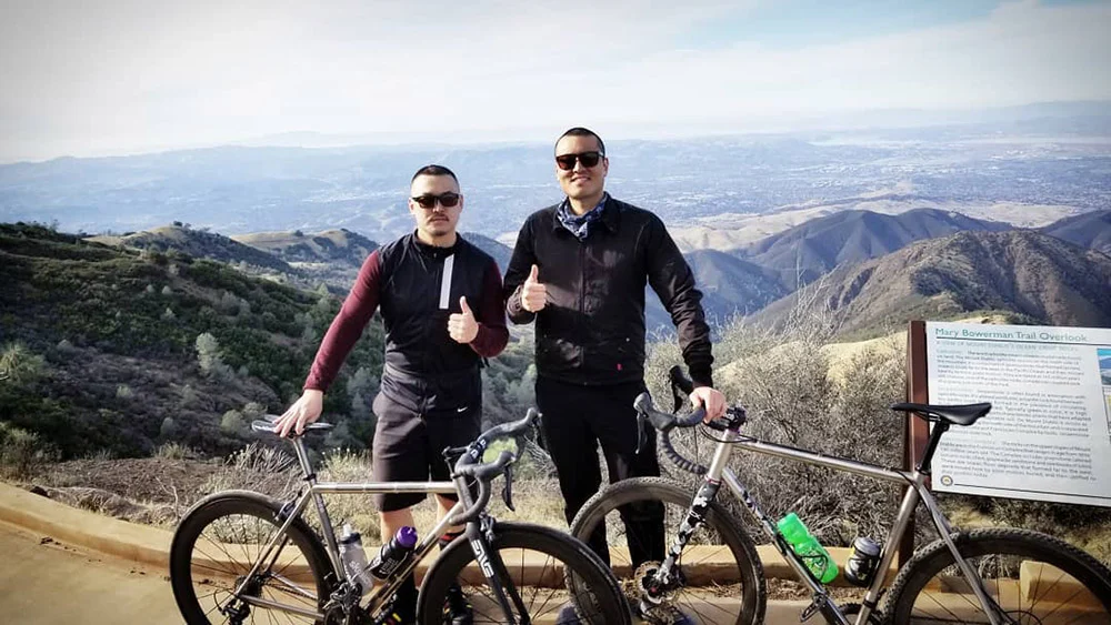 Two men give thumbs up beside mountain bikes on a dirt overlook above rolling hills.