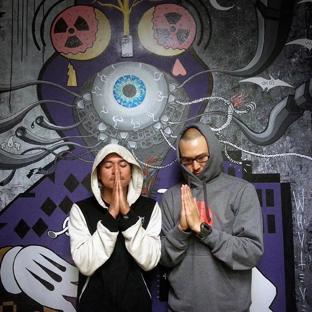 Two hooded people stand with palms pressed together in front of a purple and black mural with a large eye.