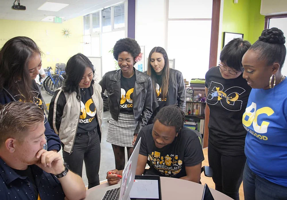 Eight people gather around a laptop in a bright room, watching a seated person use it.