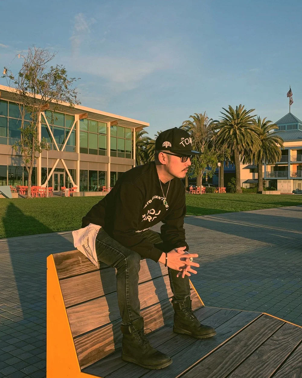 A person in a black cap and hoodie sits on a wooden bench outside modern buildings with palm trees.