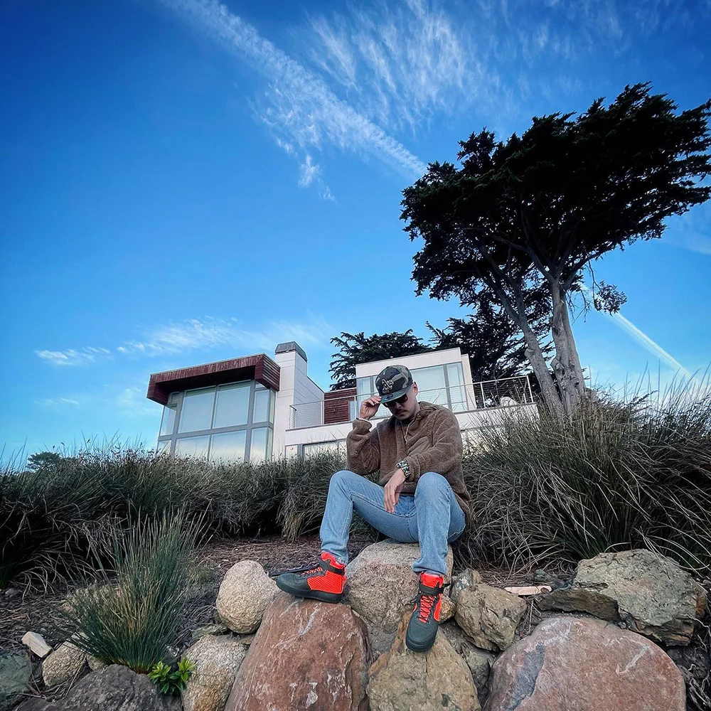 A person in a cap, brown jacket and bright-laced boots sits on rocks below a house and tall trees.