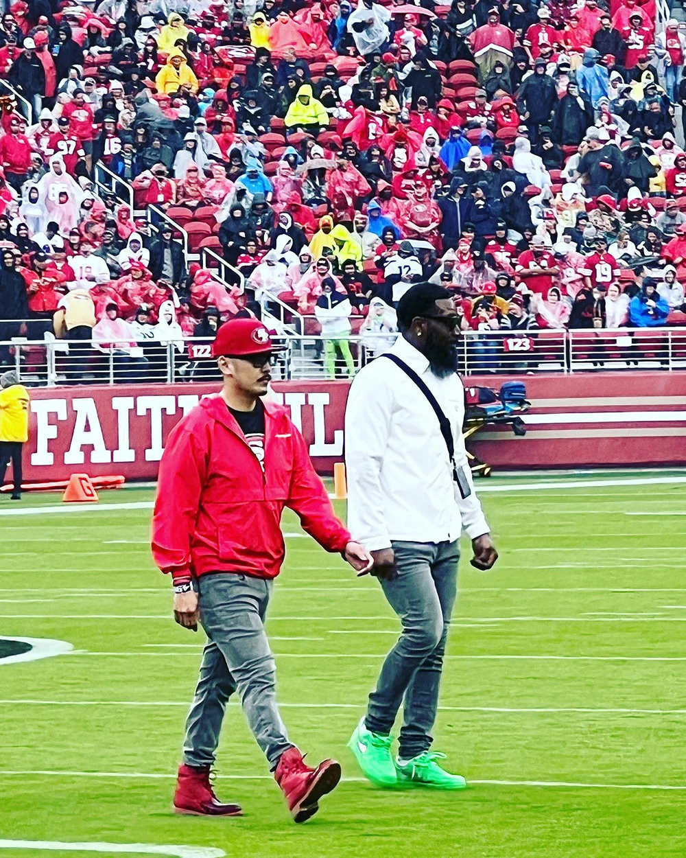 Two men in red and white jackets walk across a football field in front of packed stadium stands.