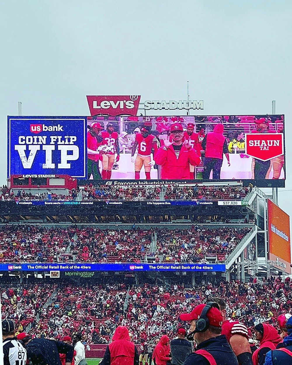 Fans fill Levi’s Stadium as a giant screen shows people on the field beside a coin flip VIP sign.