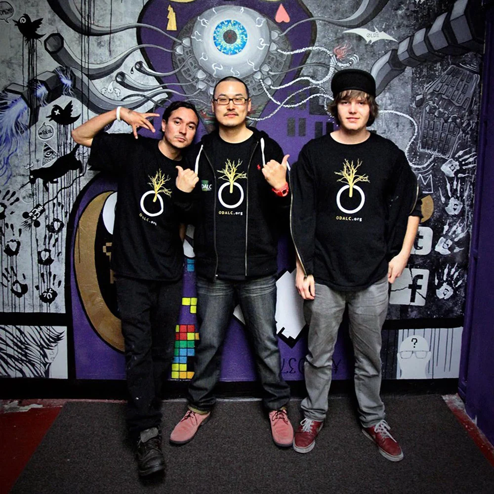 Three people in matching black shirts pose in front of a purple, black and white mural with a large eye.