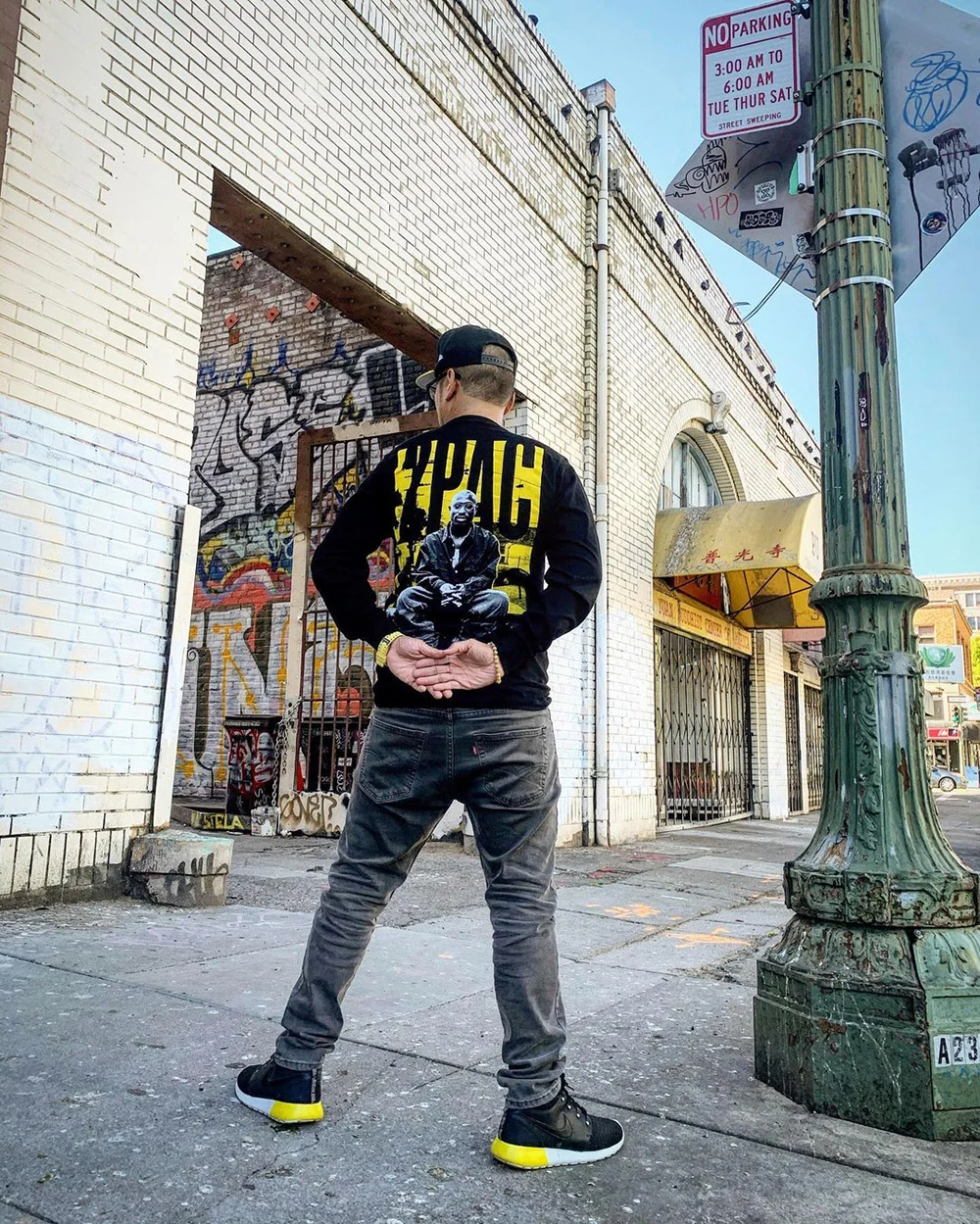 A person stands with hands behind their back on a sidewalk beside a graffiti-covered doorway and green lamppost.