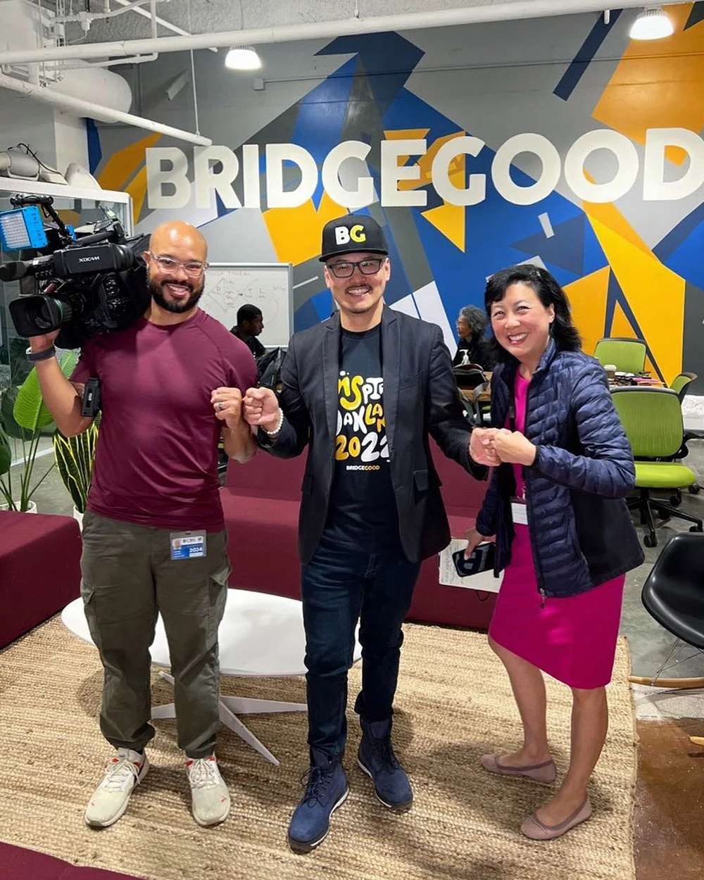 Three adults pose in a BRIDGEGOOD workspace, one holding a video camera and the others raising their fists.