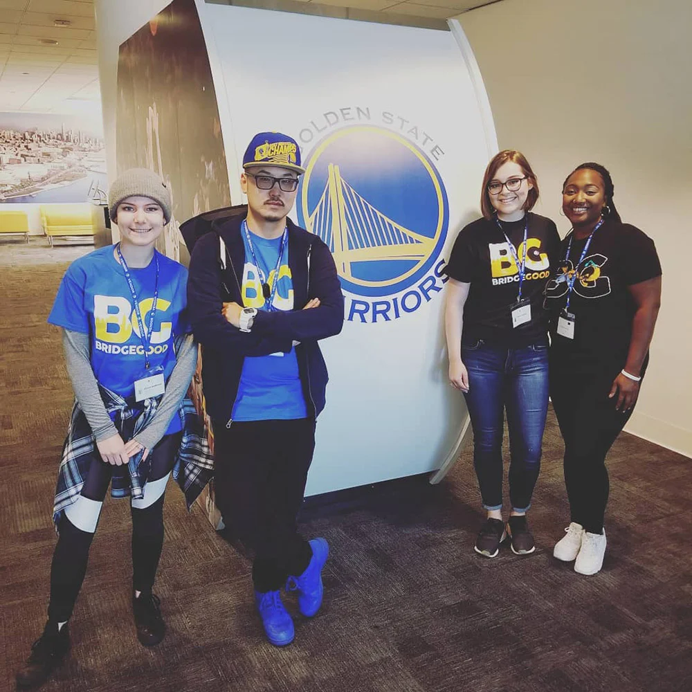Four people wearing BRIDGEGOOD shirts and badges pose beside a Golden State Warriors logo in a lobby.