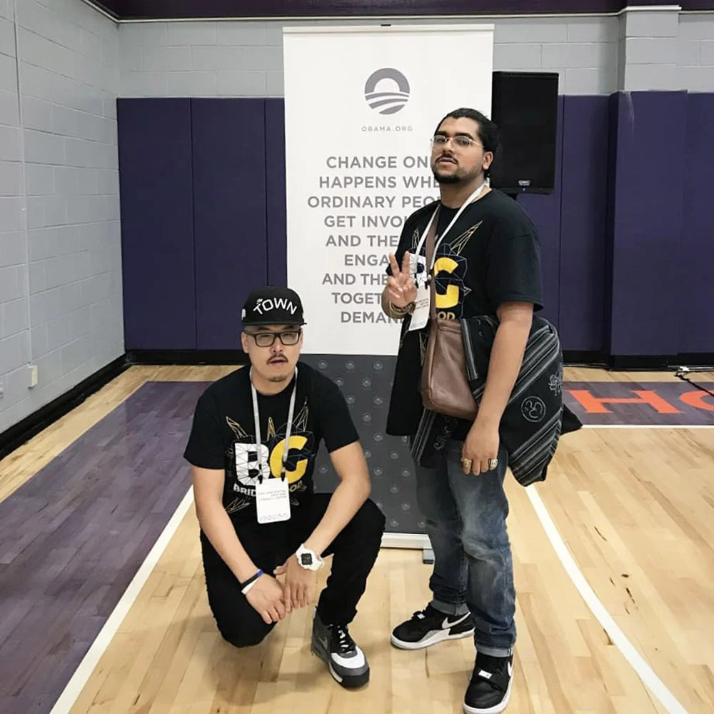 Two people wearing black shirts and event badges pose in a gym, one kneeling beside the other, near an Obama banner.