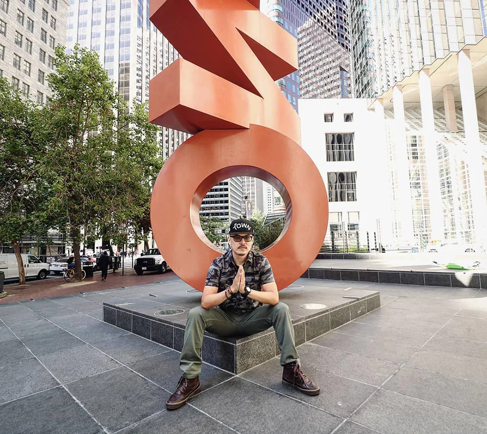 A person sits with palms together on a stone ledge beneath a large orange sculpture in a plaza surrounded by high-rises.
