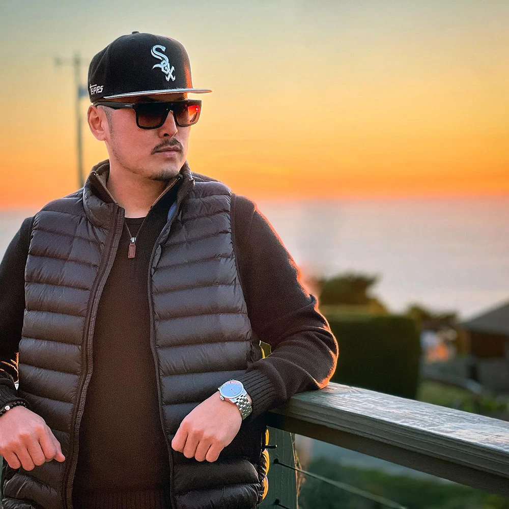 A man in a White Sox cap, sunglasses and black vest leans against a deck railing with water and sunset behind him.