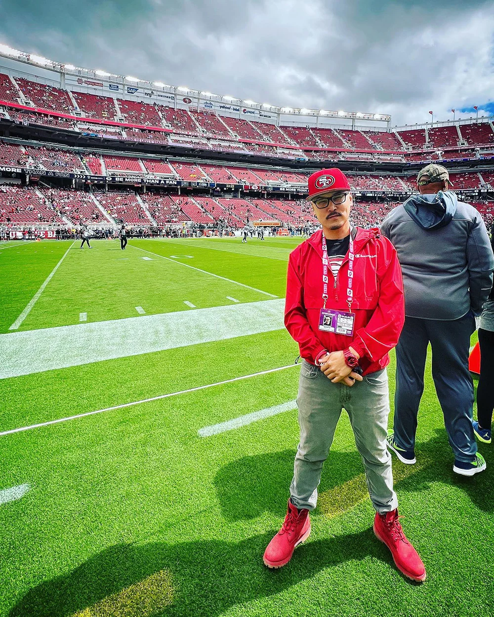 A man in red poses on a football stadium sideline, with another person facing the field and fans in the stands.