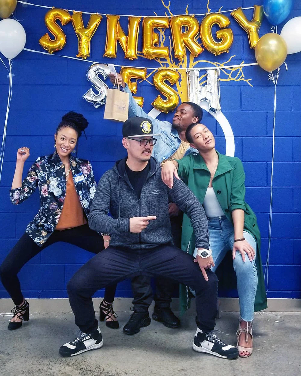 Four people strike playful poses in front of a blue wall decorated with gold and silver letter balloons.