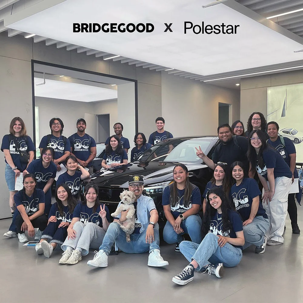 A group poses around a black car in a showroom beneath a BRIDGEGOOD x Polestar sign, with a small dog in front.