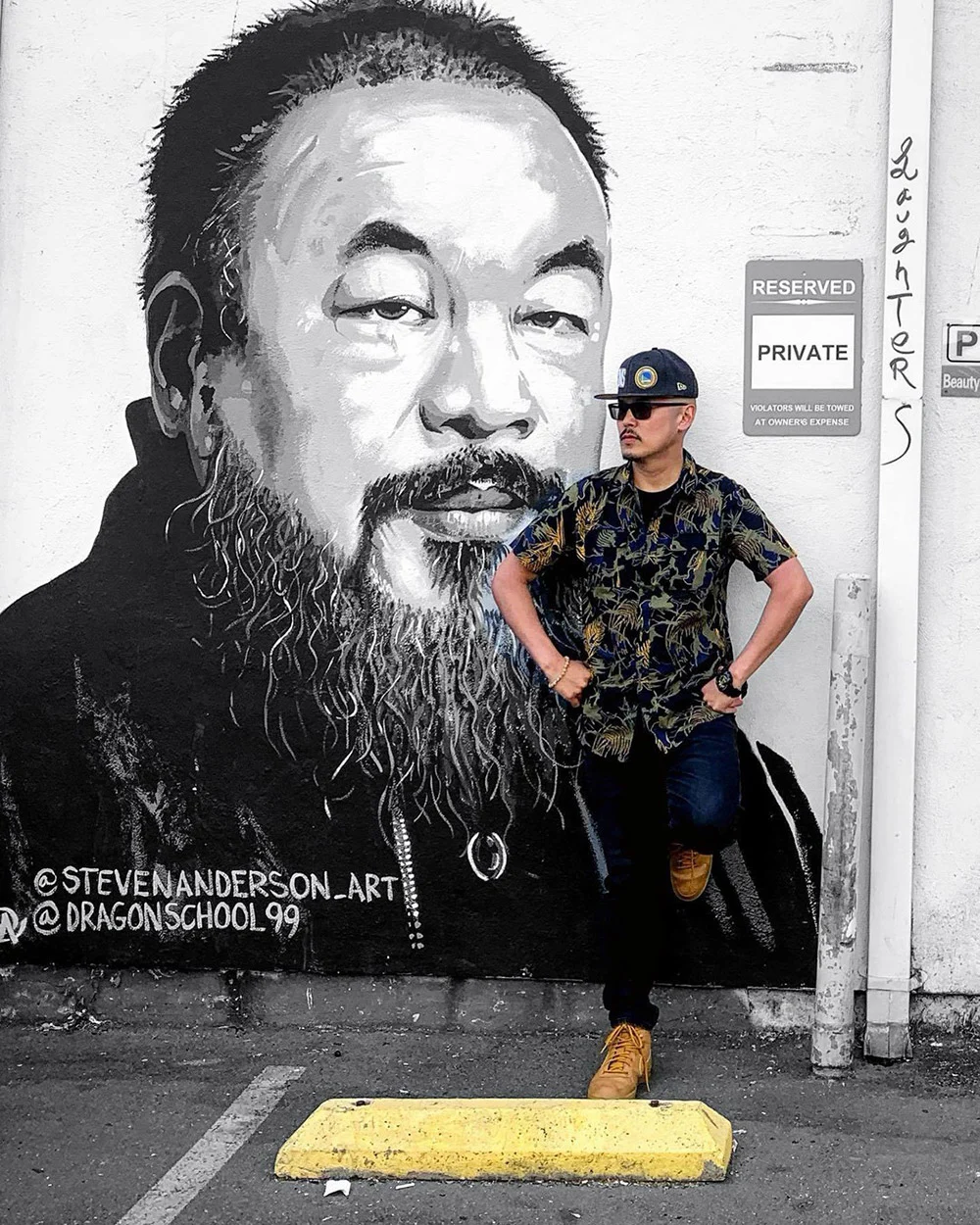 A man in a cap and sunglasses stands hands on hips beside a large black-and-white portrait mural on a wall.