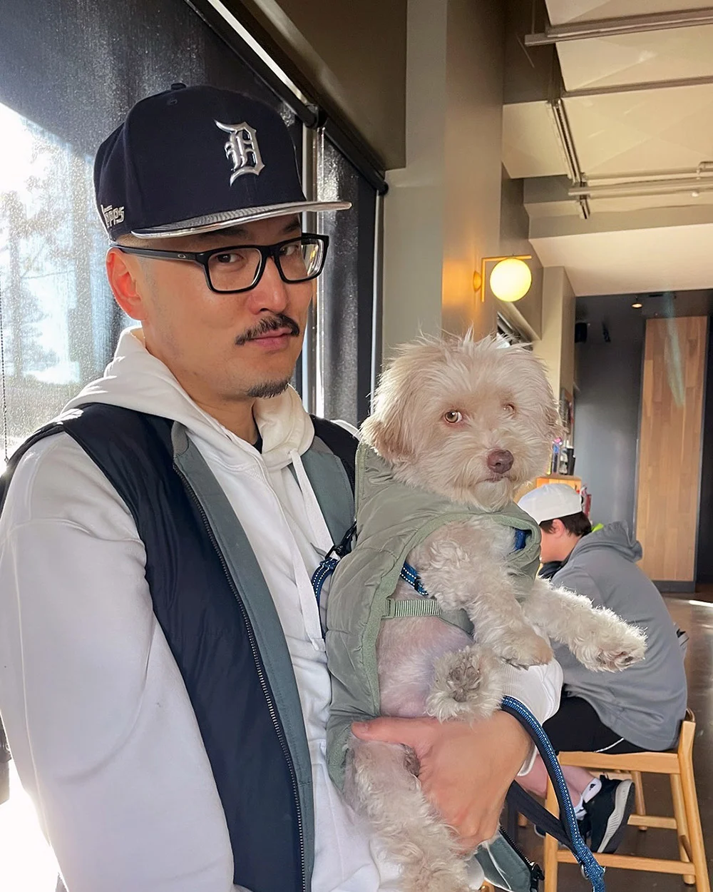 A man in glasses and a baseball cap holds a small dog in a green jacket inside a cafe.