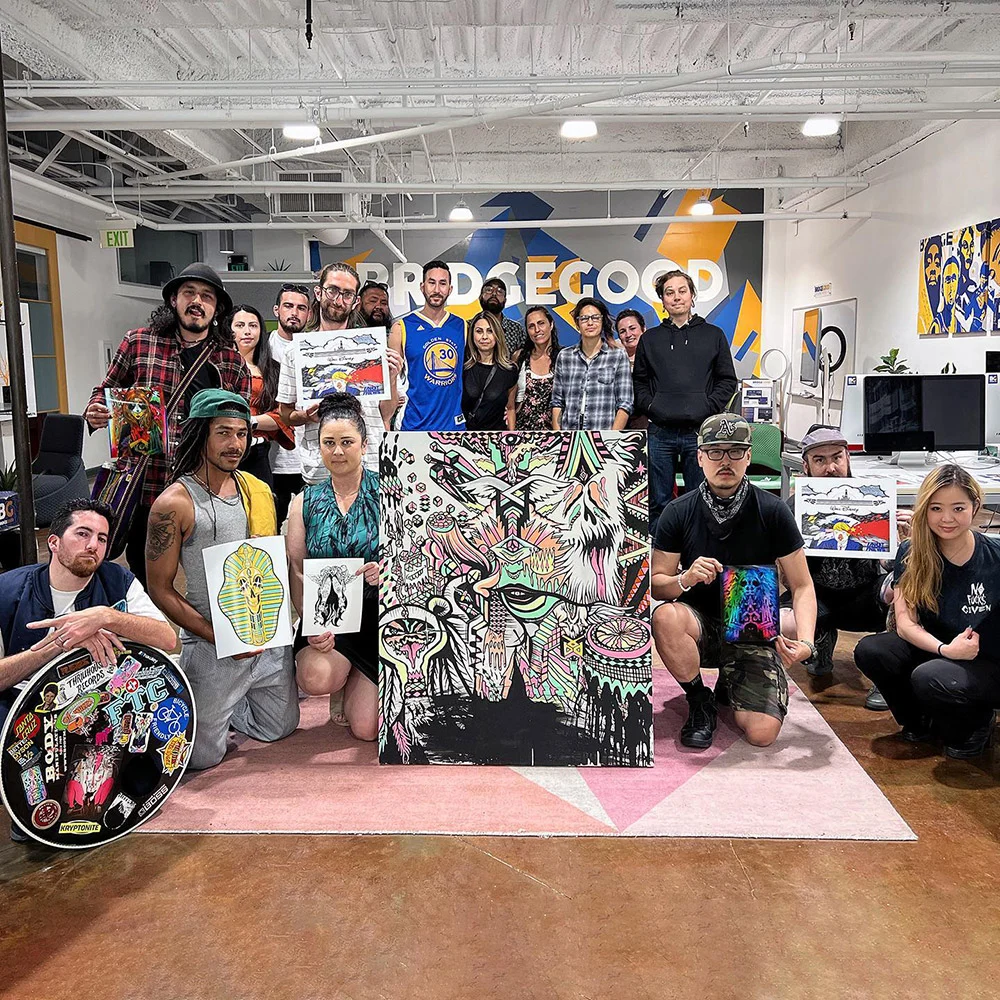 A group poses with colorful paintings and drawings in a studio beneath a large BRIDGEGOOD mural.