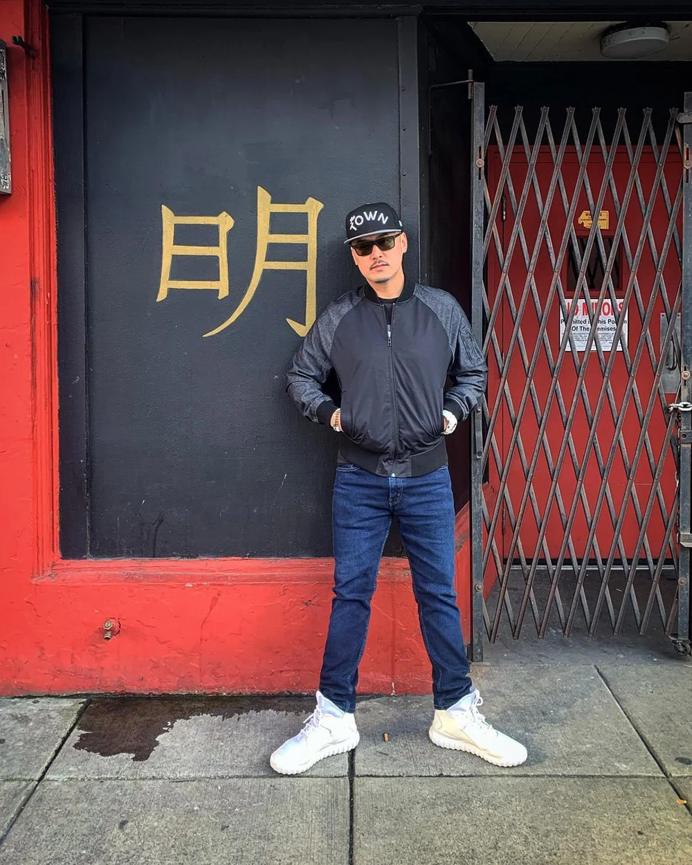 A person in a black cap, sunglasses, and jacket stands outside a red-and-black storefront beside a metal gate.