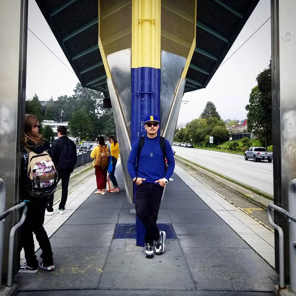 A person in blue leans against a pillar at a tram stop while others wait beside the tracks.