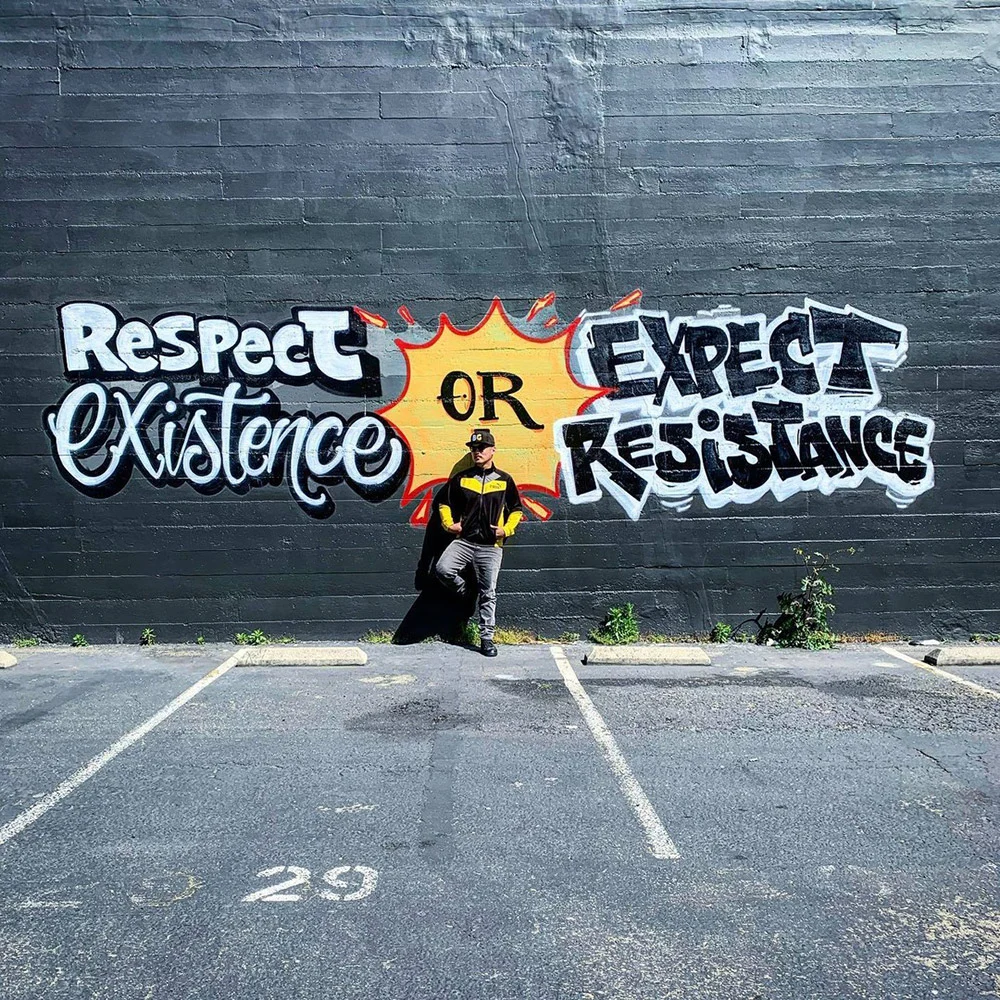 A person stands against a brick wall painted with “Respect existence or expect resistance” beside an empty parking lot.