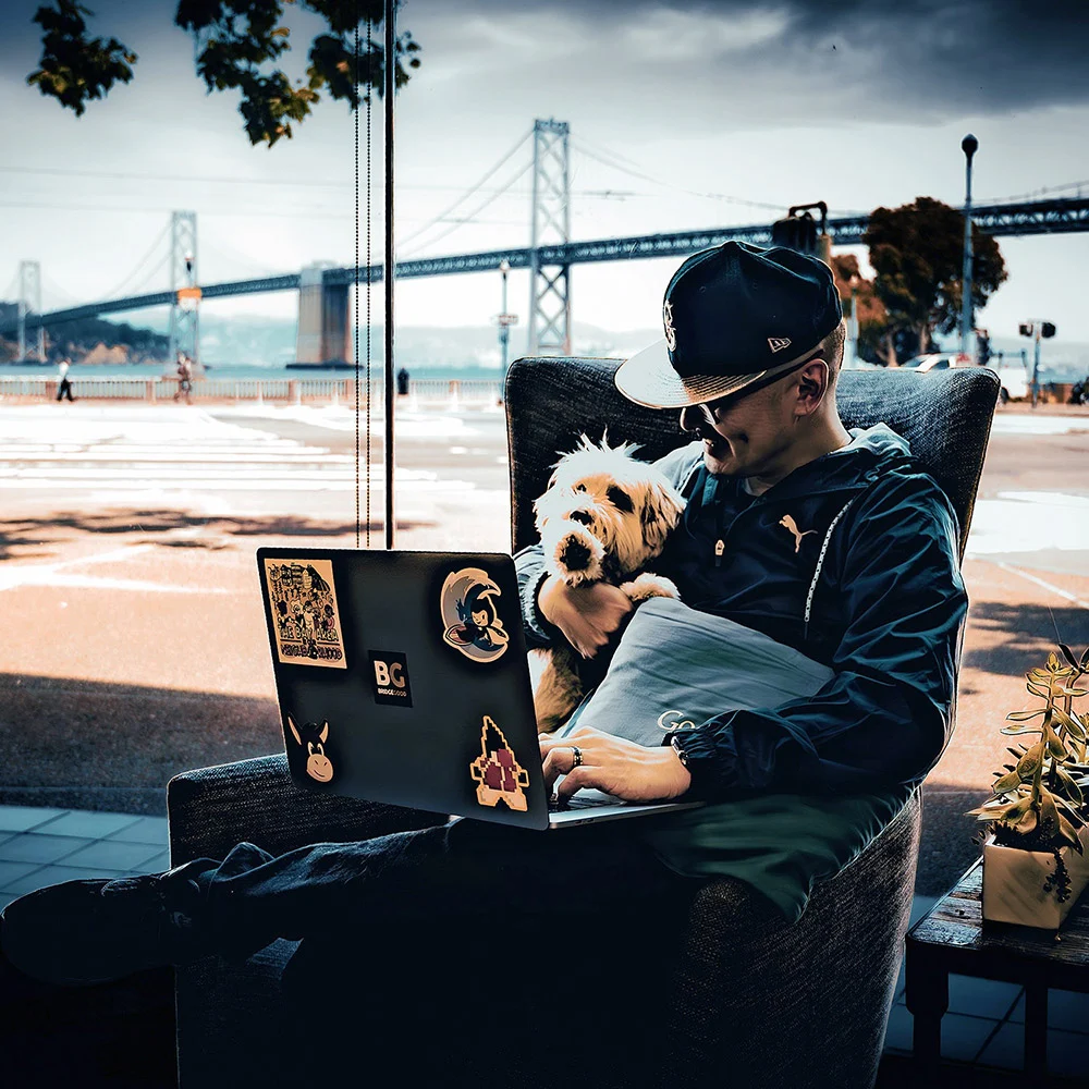 A person in a cap sits with a dog and an open laptop by a window overlooking the Bay Bridge.