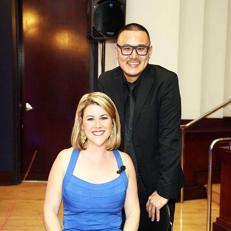 A man in a black suit and glasses stands behind a seated woman in a blue dress in a wood-floored hall.