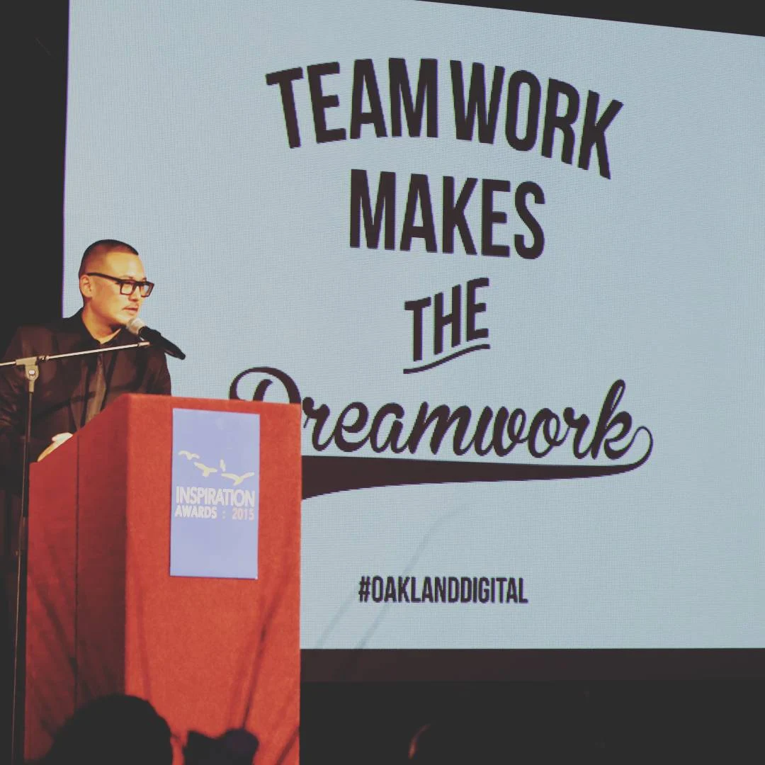 A man in glasses speaks at a red podium in front of a large screen reading Teamwork Makes the Dreamwork.