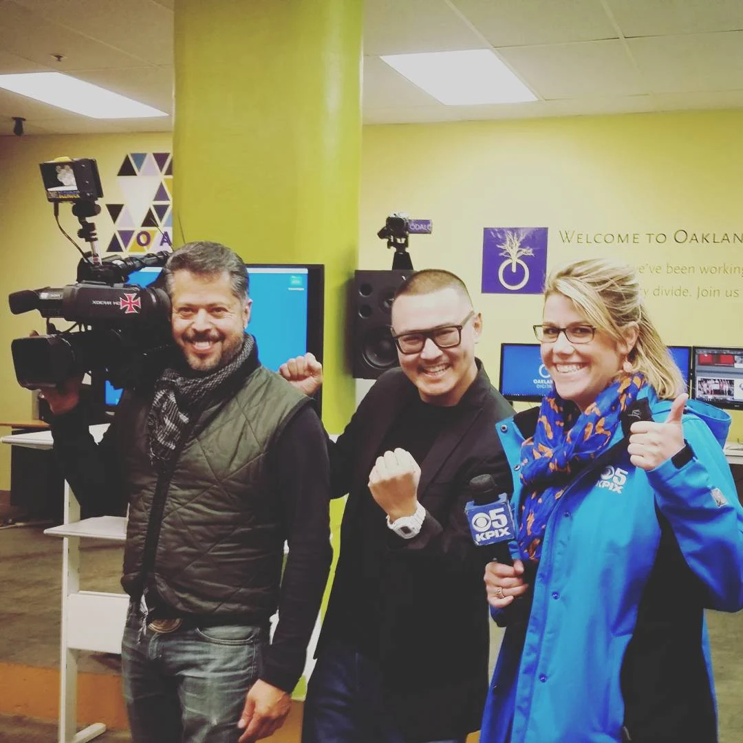 A cameraman, a man in glasses and a reporter in a blue KPIX jacket smile in a yellow office.