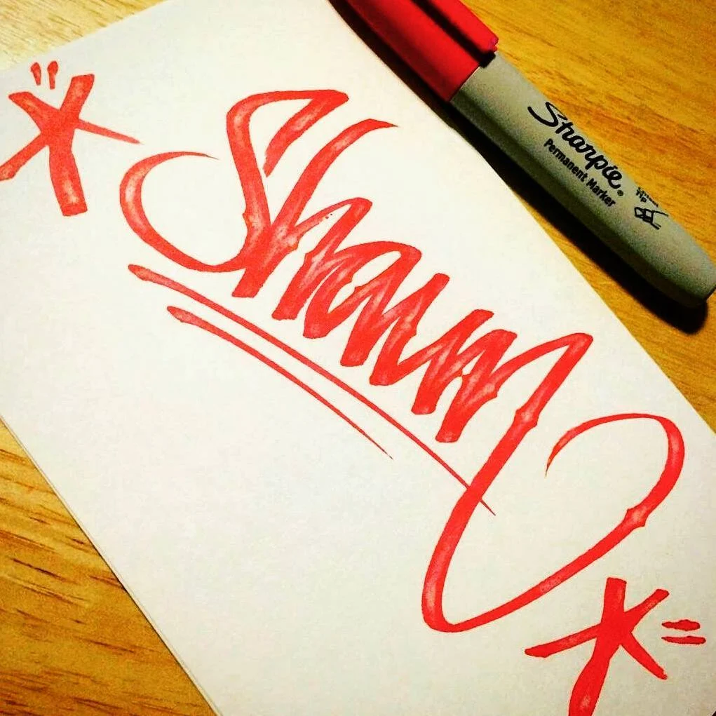 The name Shaun in red marker script on white paper, with a red Sharpie lying on a wooden surface.