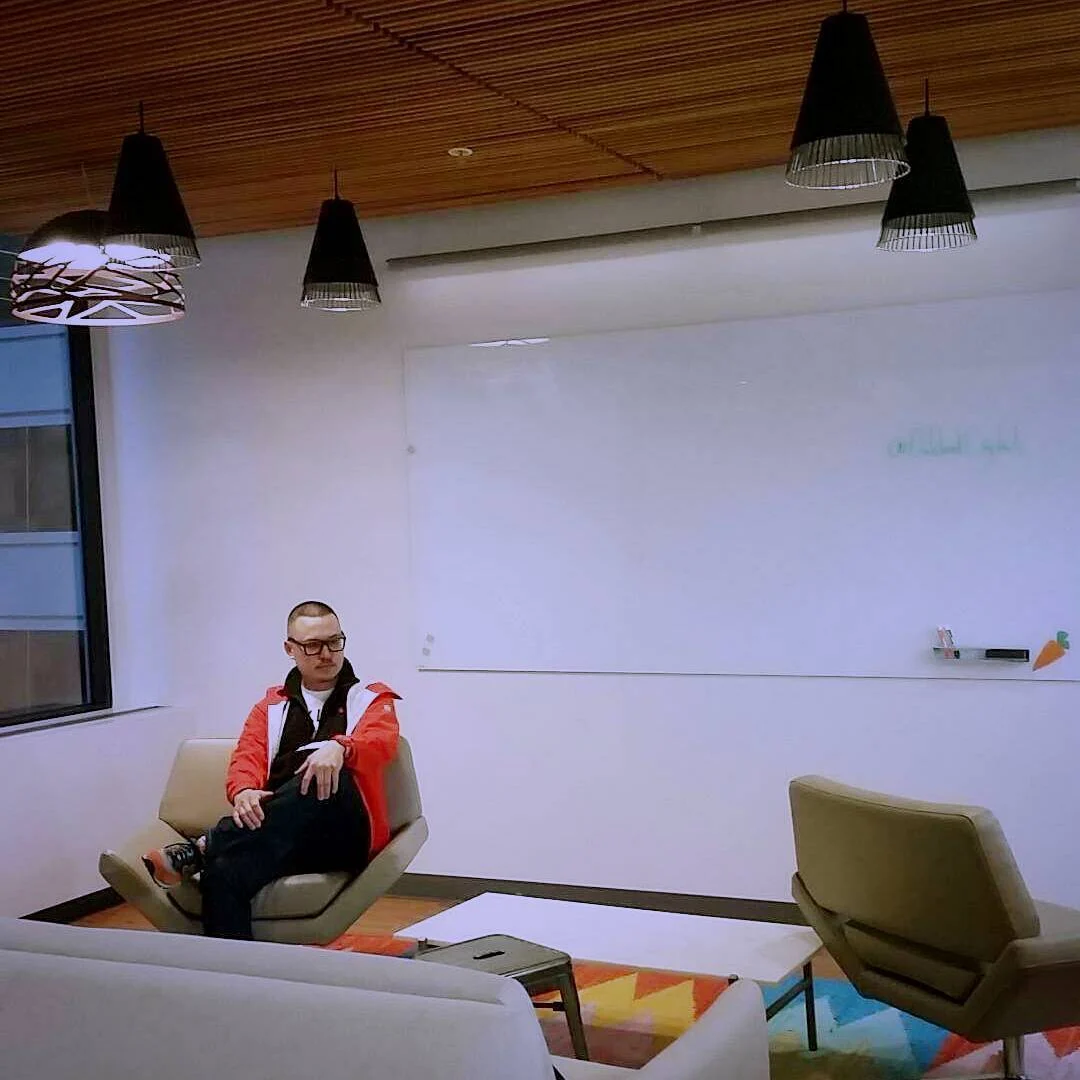A person in a red jacket and glasses sits in a beige armchair in a white lounge under black pendant lamps.