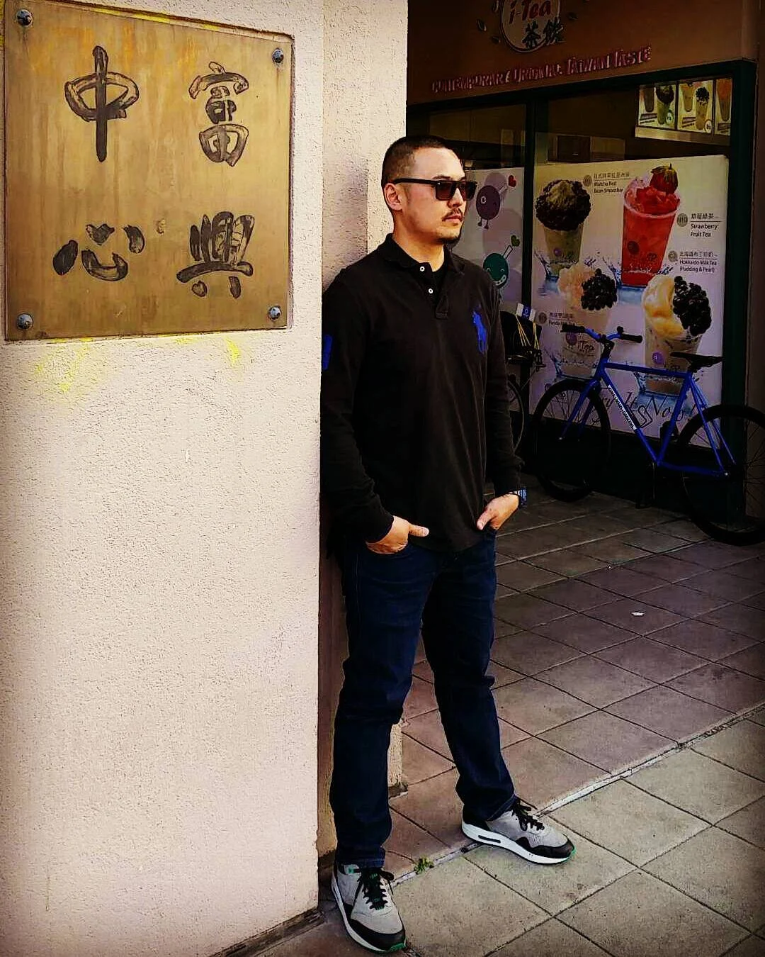 A man in sunglasses and a black polo leans against a wall with a bronze plaque, beside a boba shop and a blue bike.