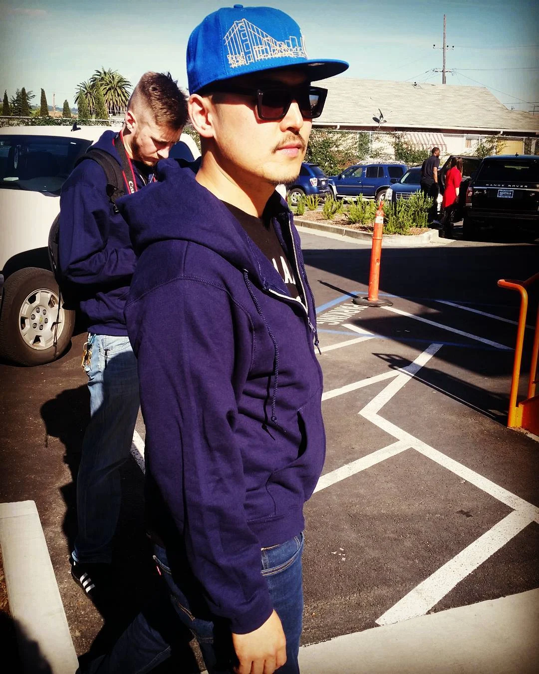 A person in a blue cap, sunglasses and navy hoodie stands in a sunny parking lot with a second person behind.