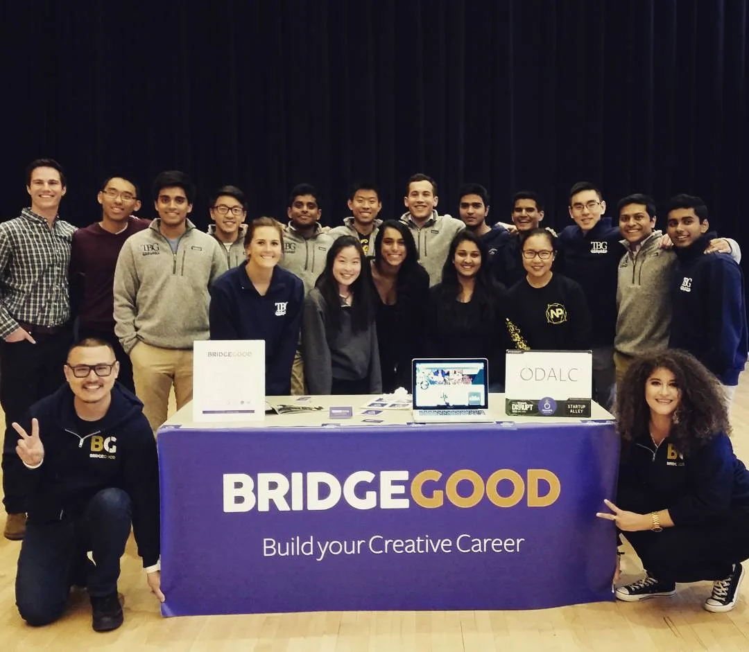 About twenty people pose behind a purple BRIDGEGOOD table banner that reads Build your Creative Career on a gym floor.